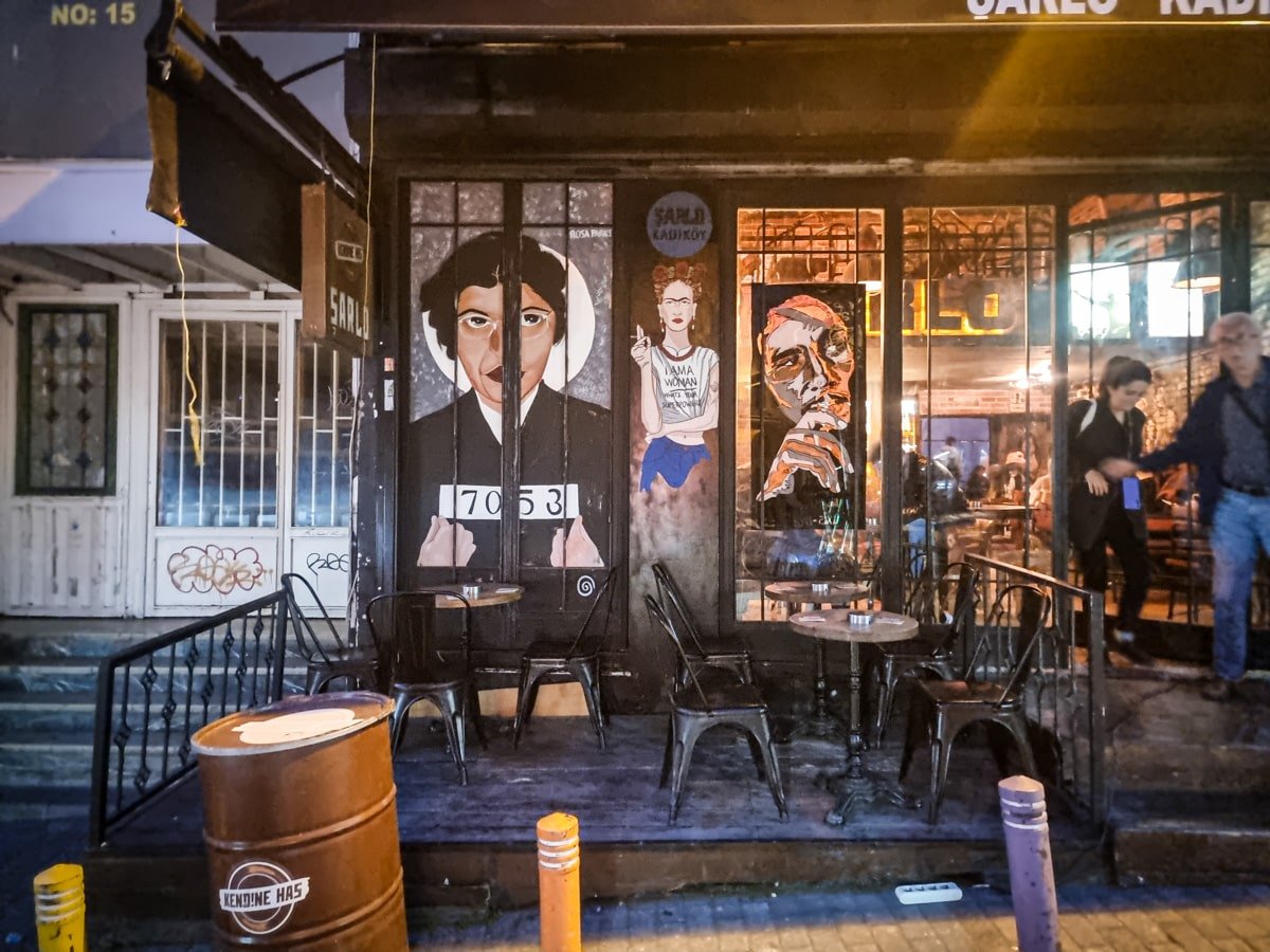 Outdoor street cafe at night with murals of three figures on its exterior wall and several tables and chairs in front. A barrel with a beverage logo is in the foreground, and two people stand nearby, capturing a scene straight out of *A Guide To Kadikoy Istanbul*.