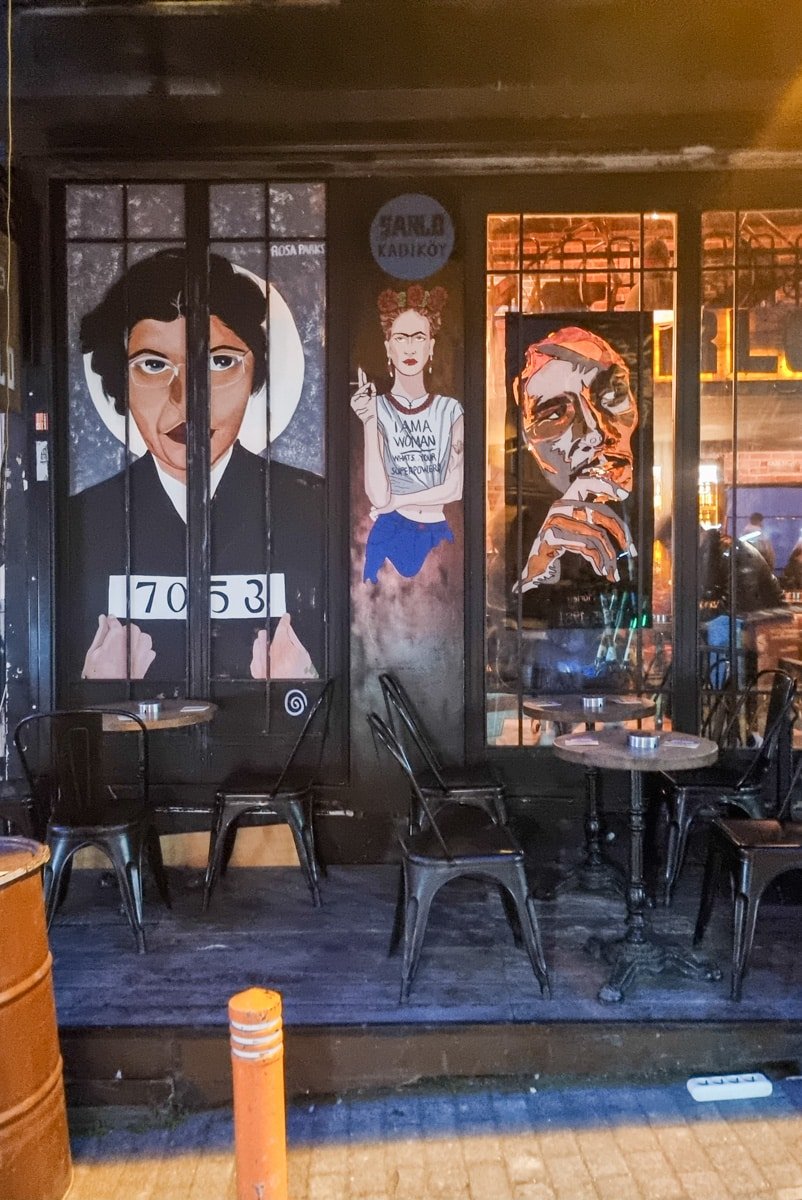 A street art mural featuring three distinct portraits graces a cafe's exterior wall in Kadikoy, Istanbul, with empty black chairs and tables in front.
