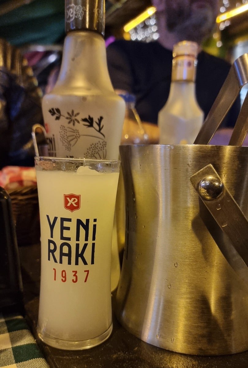 A bottle of Yeni Rakı, a glass with "Yeni Rakı 1937" printed on it, and a metal ice bucket on a table—perfect elements to begin A Guide To Kadikoy Istanbul.