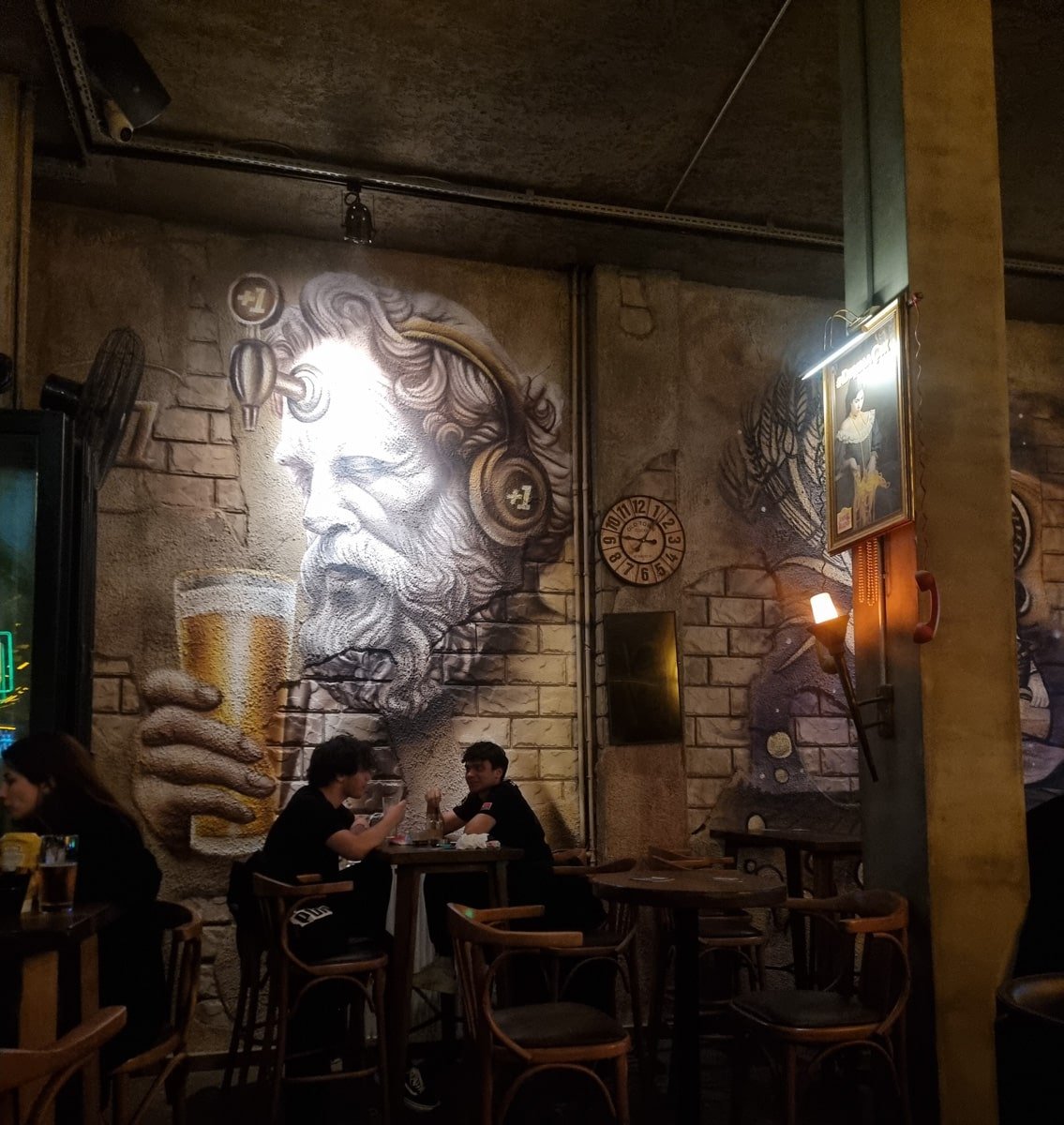 Two people sit at a wooden table in a dimly lit pub with a large mural of a bearded man holding a beer on the wall behind them, reminiscent of scenes from *A Guide to Kadikoy Istanbul*.