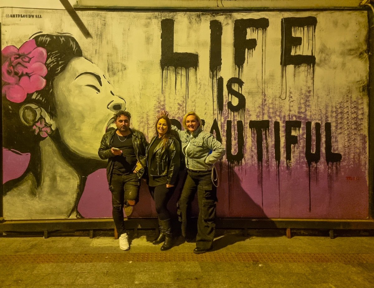Three people stand in front of a mural with the text "LIFE IS BEAUTIFUL" painted on it, reminiscent of the vibrant street art you'll find in A Guide To Kadikoy Istanbul. The mural features a side profile of a woman with flowers in her hair, capturing the essence of urban creativity.