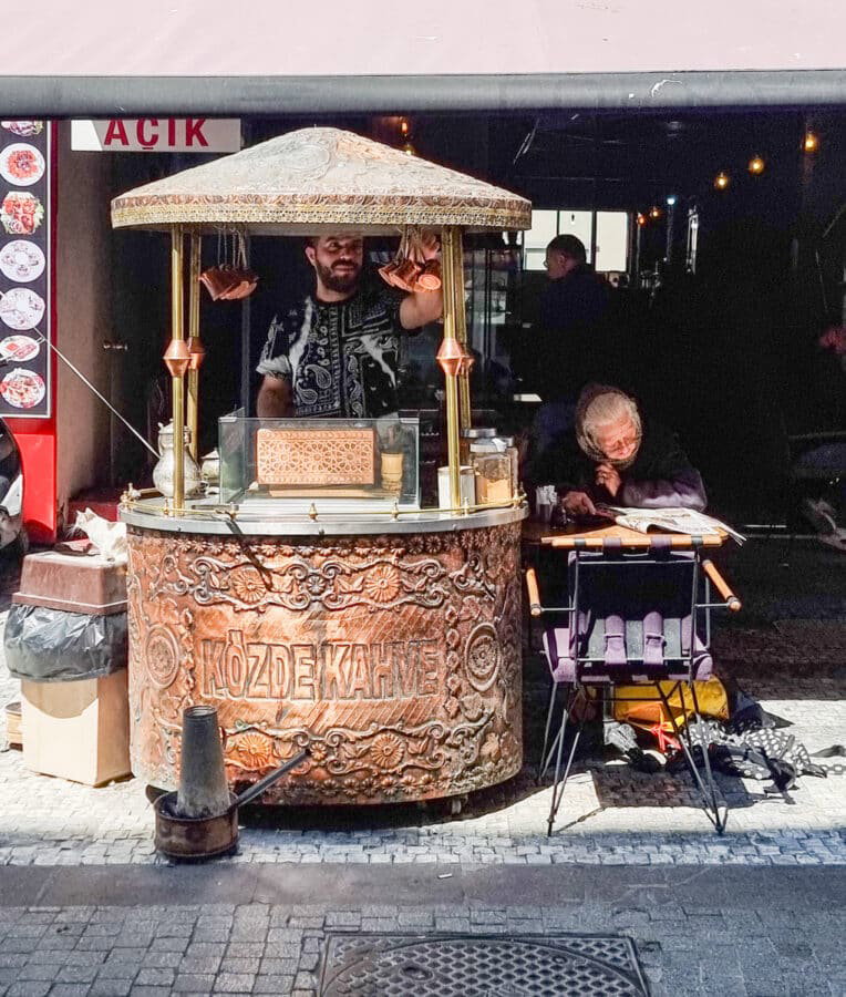A vendor stands behind a copper coffee cart labeled "Közde Kahve" under a canopy. A seated elderly man reads a book nearby on the cobblestone street, reminiscent of scenes described in *A Guide to Kadikoy Istanbul*.