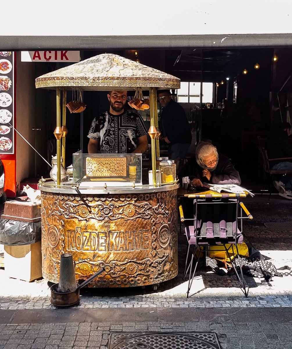 A man stands behind an ornate, cylindrical coffee stand labeled "Közde Kahve" while an elderly woman sits nearby under an umbrella on a street in Kadikoy, Istanbul.