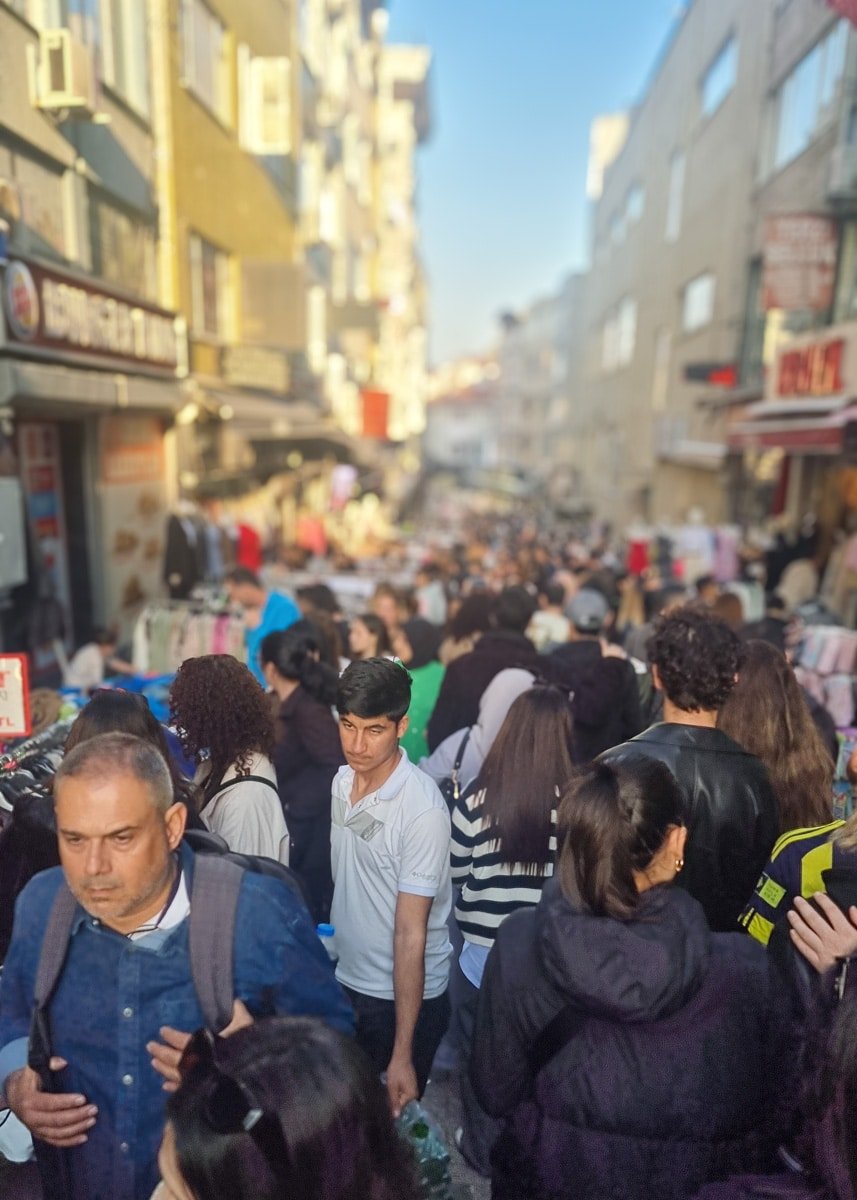 A crowded street market with many people walking through, surrounded by buildings and various stalls. The scene is bustling under clear daylight, much like a snapshot from A Guide to Kadikoy Istanbul.