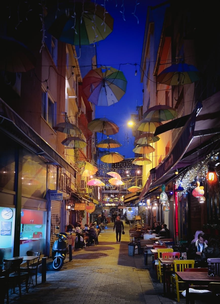 A Guide to Kadikoy Istanbul: Narrow streets at night, decked with colorful hanging umbrellas and dim lighting, invite you to enjoy the outdoor seating while people casually stroll by.
