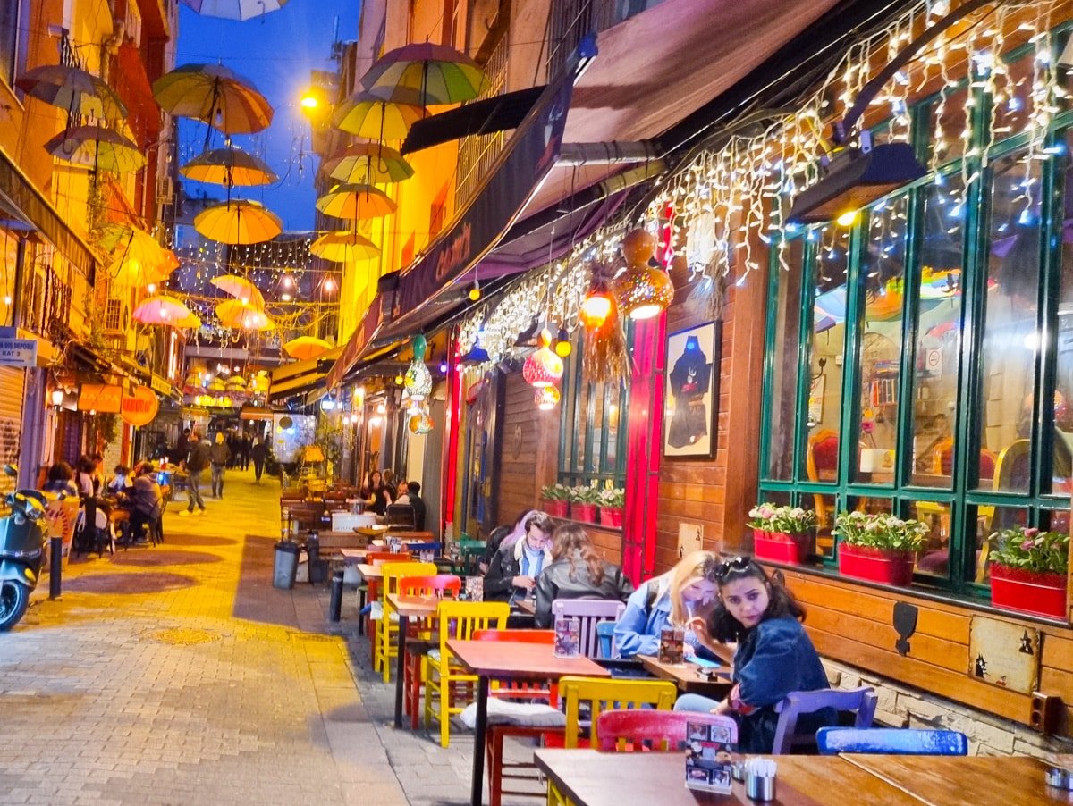 A lively outdoor cafe on a narrow street in Kadikoy, Istanbul, decorated with colorful umbrellas, string lights, and potted plants. People are dining at tables along the sidewalk under the evening sky.
