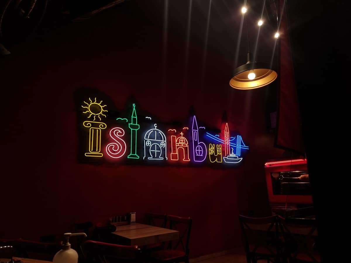 A neon sign reading "Istanbul" with colorful depictions of the city's landmarks is displayed on a dark wall, reminiscent of *A Guide To Kadikoy Istanbul*. The room is dimly lit with a hanging light fixture and empty tables beneath the sign.