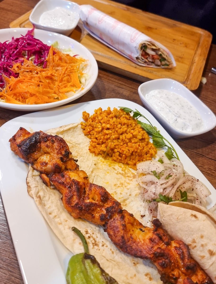 A Guide To Kadikoy Istanbul: A plate with grilled kebab on flatbread, bulgur, green pepper, sliced onions, and a second plate with a wrap and salads. A bowl of white sauce is beside the wrap.