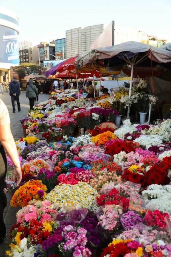 Outdoor flower market with colorful blooms displayed on tables under red umbrellas, with people walking by in a city setting.