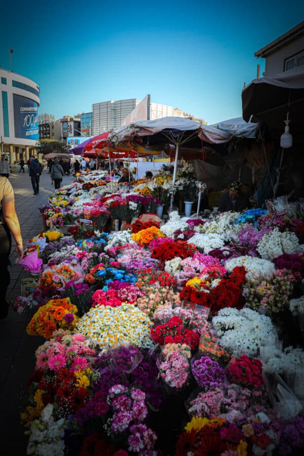 Outdoor flower market with vibrant bouquets displayed under tents, bustling street and buildings in the background.