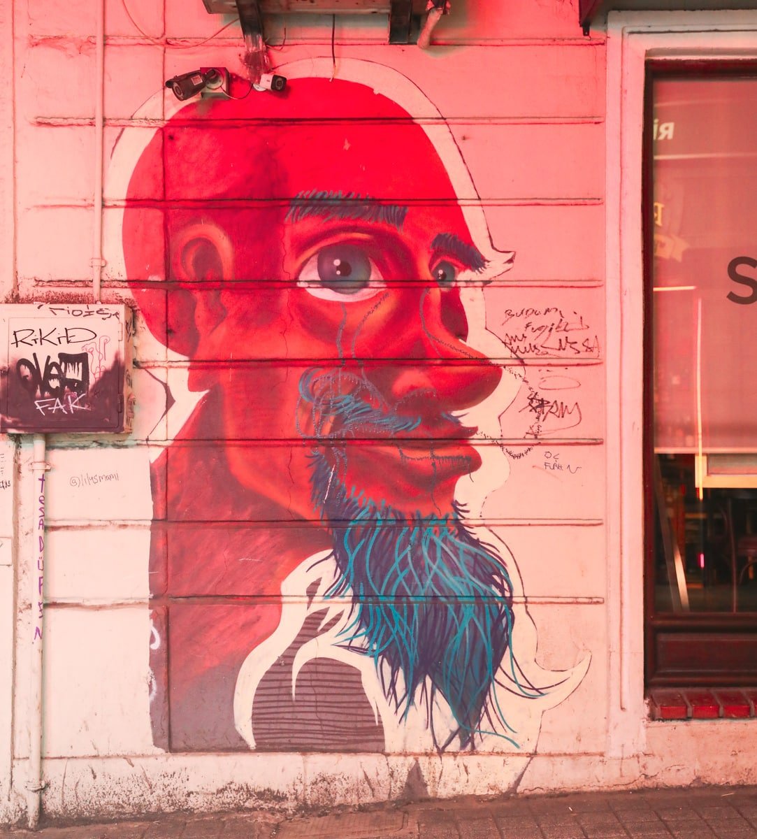 A colorful mural of a bearded man's face painted on an urban wall, with light graffiti and tags surrounding it.