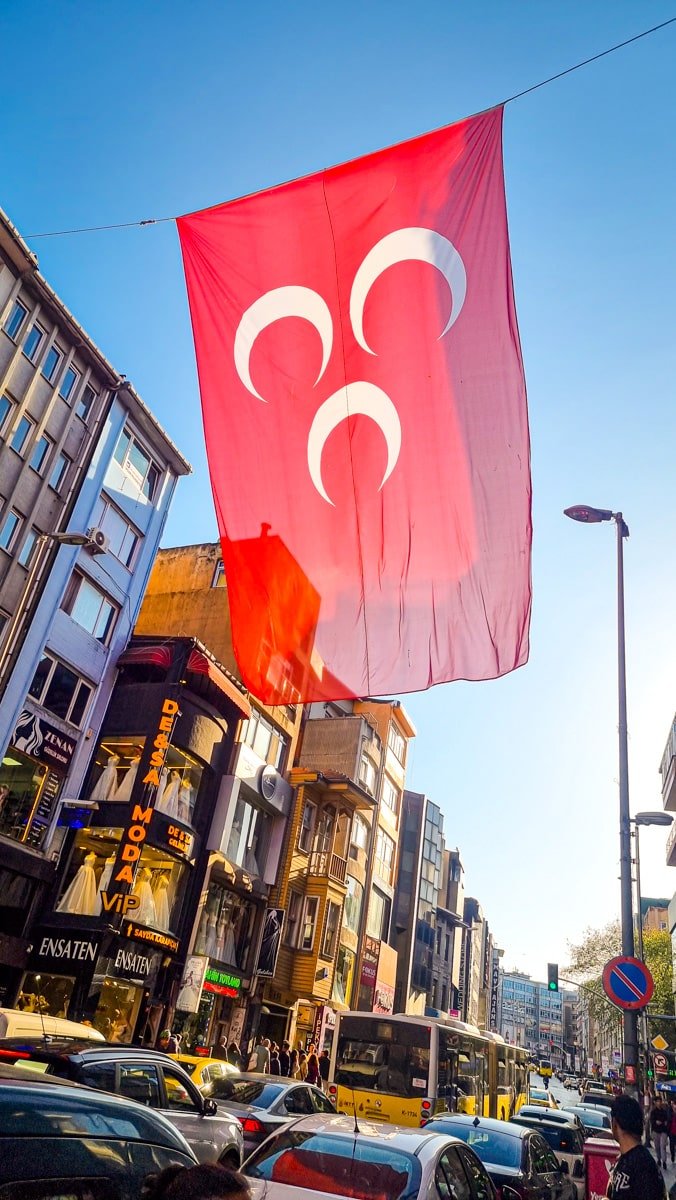 A large Turkish flag with a white crescent and star hangs over a busy street lined with shops and traffic in bright daylight.