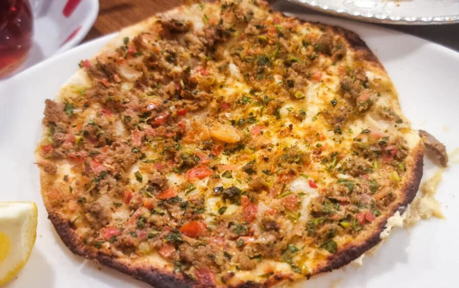 A close-up image of a Lahmacun, a Turkish flatbread topped with minced meat, vegetables, and herbs, partially eaten, on a wooden table.