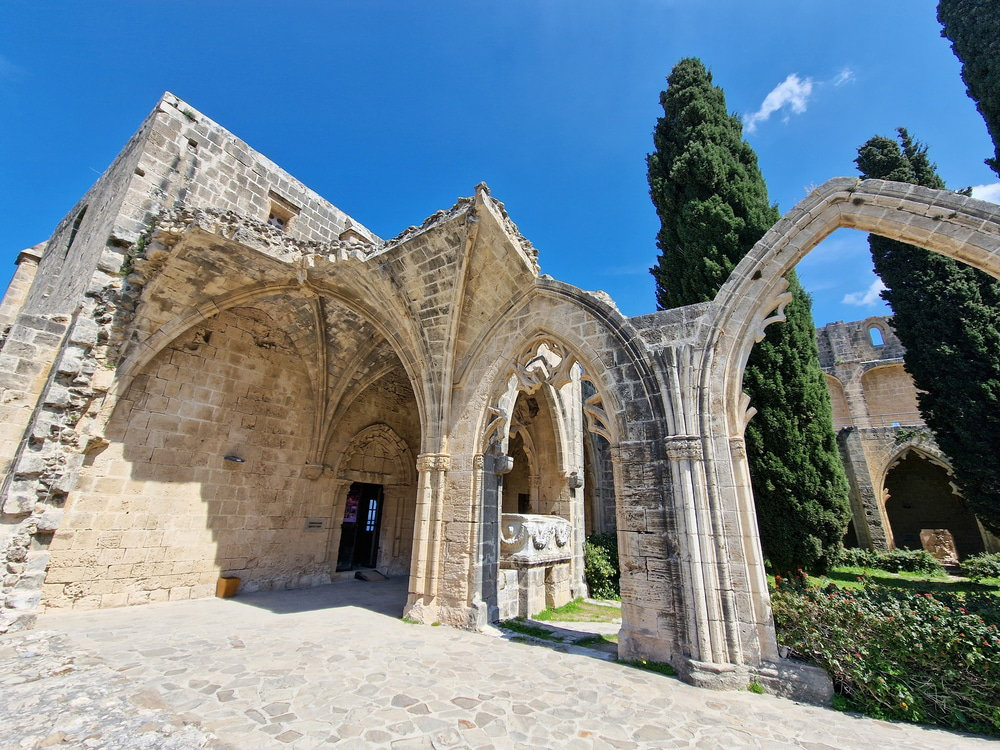 A stone medieval building with arched columns and a partly open roof stands under a bright blue sky surrounded by cypress trees. The structure features intricate stonework and a cobblestone pathway, making it one of the captivating North Cyprus attractions you shouldn't miss.