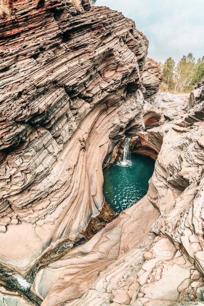 Natural rock formations surround a small pool with clear blue water, fed by a waterfall. The terrain is rugged with striated rock surfaces, and sparse vegetation is visible in the background. One of Australia's finest natural swimming spots Karijini Rock Pool