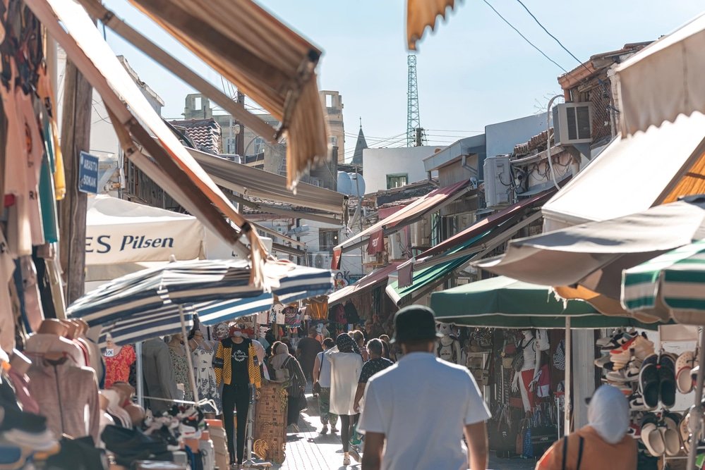 A bustling outdoor market in North Cyprus with people walking between stalls selling various goods under awnings and umbrellas on a sunny day—a must-see spot featured in every travel guide for its vibrant atmosphere and local charm, adding to the list of things to do while visiting.