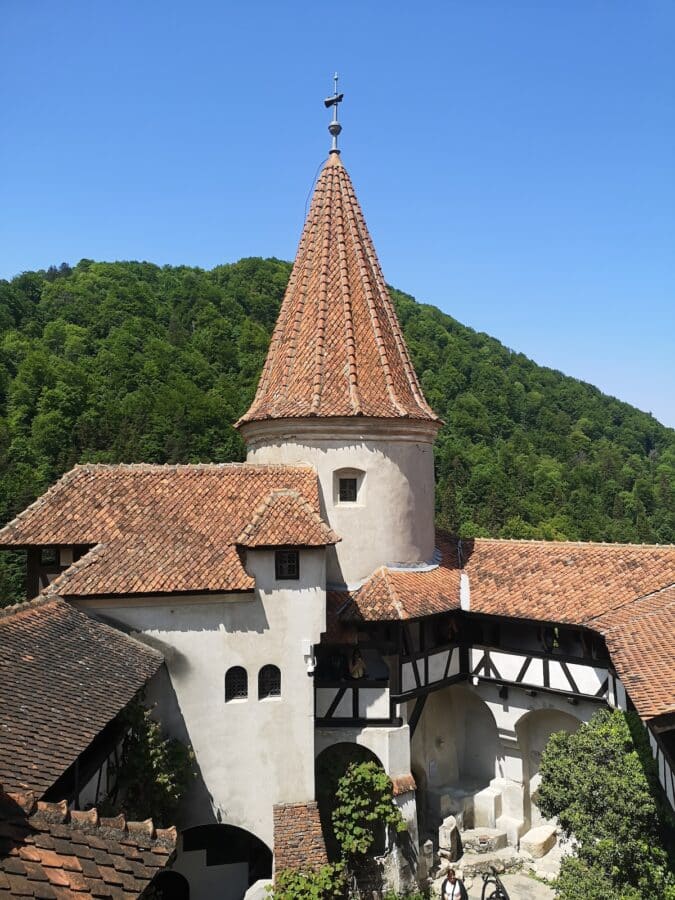 A medieval building with a conical tower topped with a cross, surrounded by lush green hills under a clear blue sky. The structure has red-tiled roofs and white walls, reminiscent of tales from Myths and Legends—a perfect stop on any Romania Road Trip.