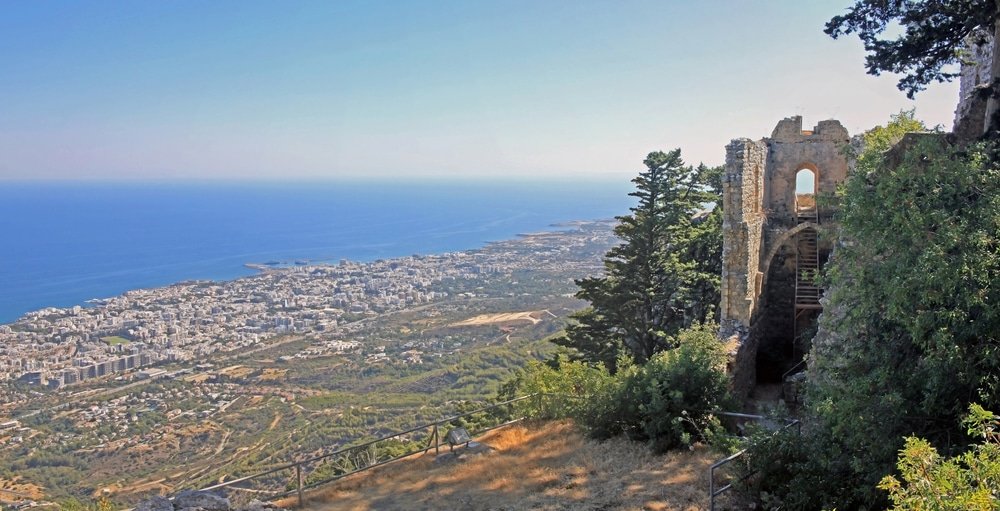 Ready for a breathtaking adventure? Enjoy the stunning view of North Cyprus' coastal city from the vantage point next to ancient stone ruins, with a clear blue sea in the background and lush green vegetation in the foreground. Discover endless things to do in this picturesque paradise.