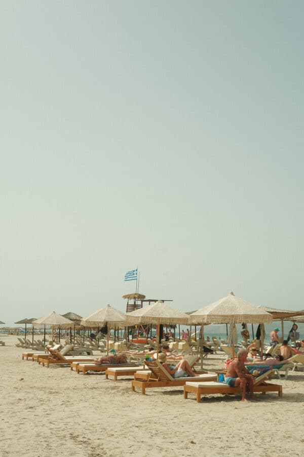 Beachgoers relax on wooden sun loungers under straw umbrellas. A Greek flag is visible in the background near the lifeguard hut on this beautiful day in Kos. The sky is clear, and the sea is visible in the distance, capturing the essence of the Greek Islands.