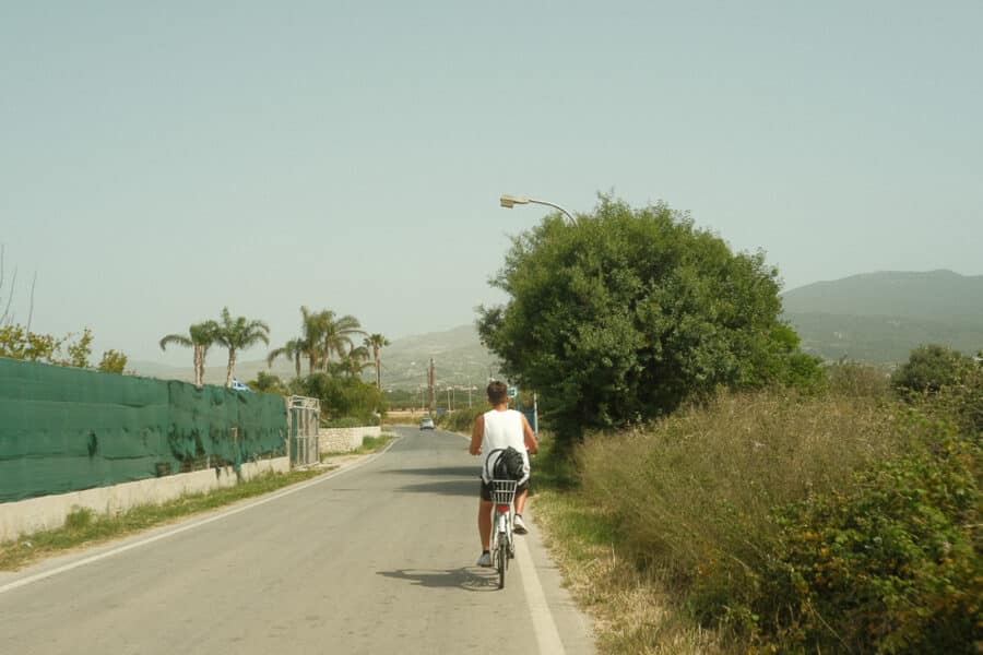 A person rides a bicycle down a paved road bordered by greenery, with distant hills and a few palm trees visible in the background, reminiscent of the scenic beauty found on the Greek Islands like Rhodes and Kos.