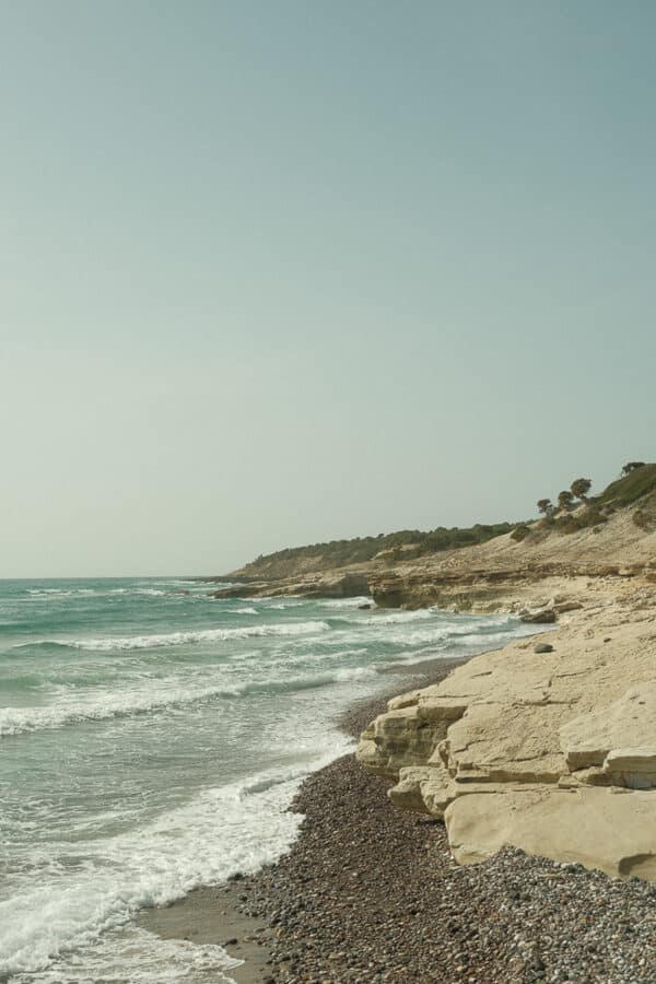 A coastal shoreline on Kos with rocky cliffs and a pebble beach, waves crashing against the shore. Sparse greenery is visible on the cliff tops under a clear sky.