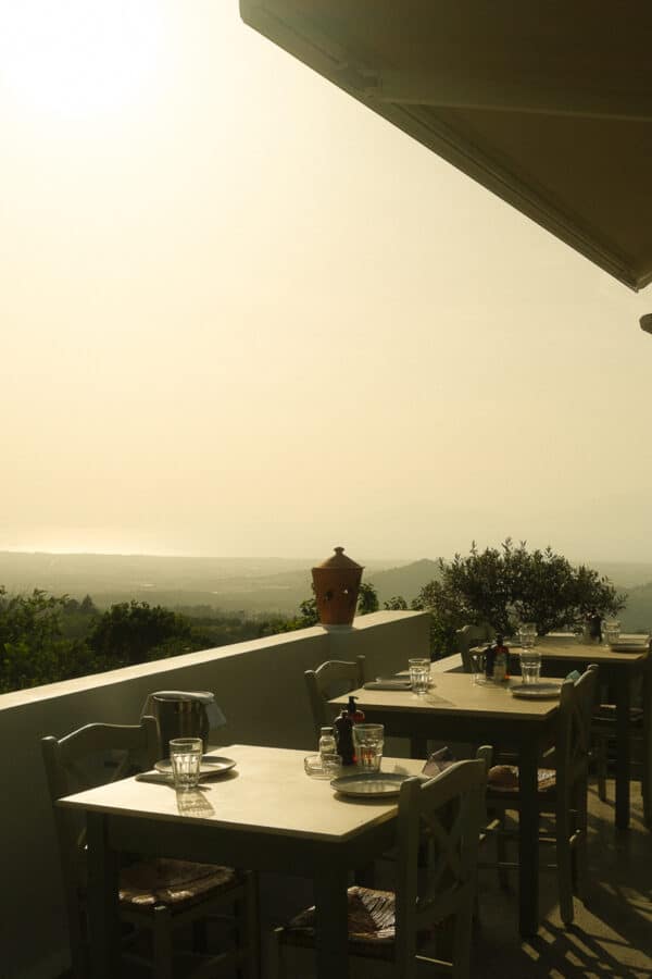 Outdoor terrace with several empty tables and chairs, overlooking a hazy, scenic landscape reminiscent of the Greek Islands, with trees and distant mountains. Daylight gives a warm, atmospheric glow to the setting.