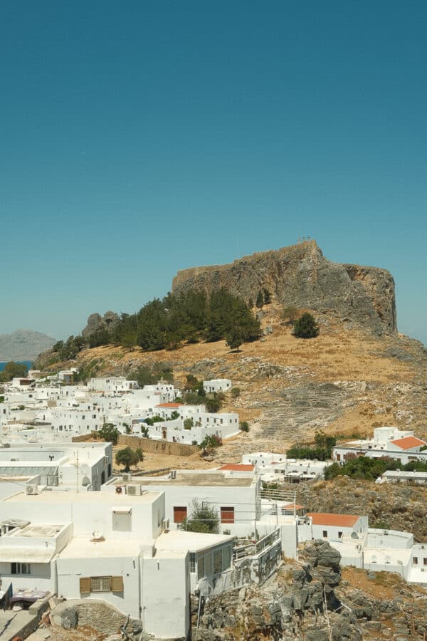 A rocky hill topped with ancient ruins overlooks a densely packed cluster of white buildings under a clear blue sky, evoking the timeless beauty reminiscent of the Greek Islands.