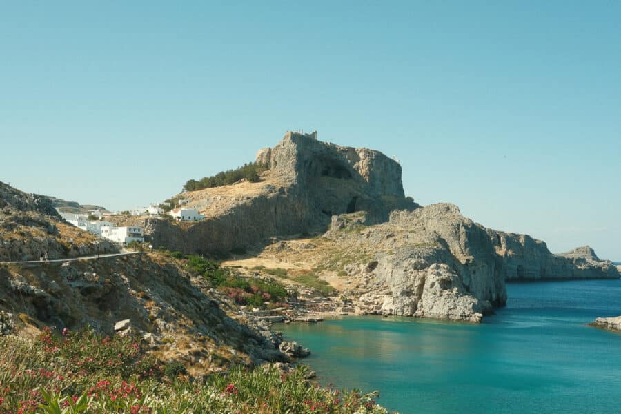 A coastal landscape with rocky cliffs, clear blue water, and white buildings scattered on the hillside, reminiscent of the beauty found in the Greek Islands. A fortress is visible atop the highest cliff under a clear blue sky.