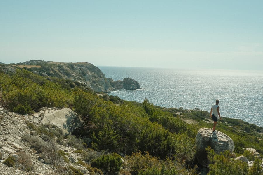 A person stands on a large rock overlooking a hilly coastal landscape with green shrubs and the vast blue ocean of Rhodes under a clear sky.