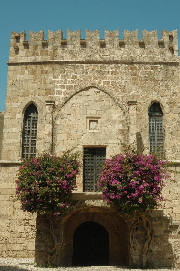 A historic stone building with arched windows and a main entrance, adorned with climbing purple flowering plants flanking the doorway, reminiscent of the charming architecture found in Kos and Rhodes of the Greek Islands.