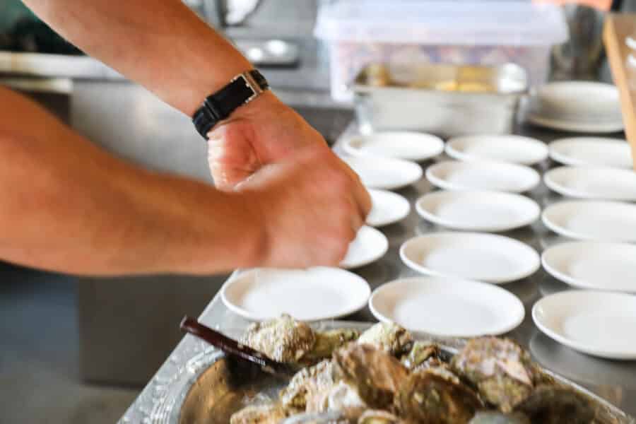 Hands shucking oysters at a kitchen counter, with several small white plates in the background, reminiscent of a delightful Bota Sare Ston Oyster Day Trip.