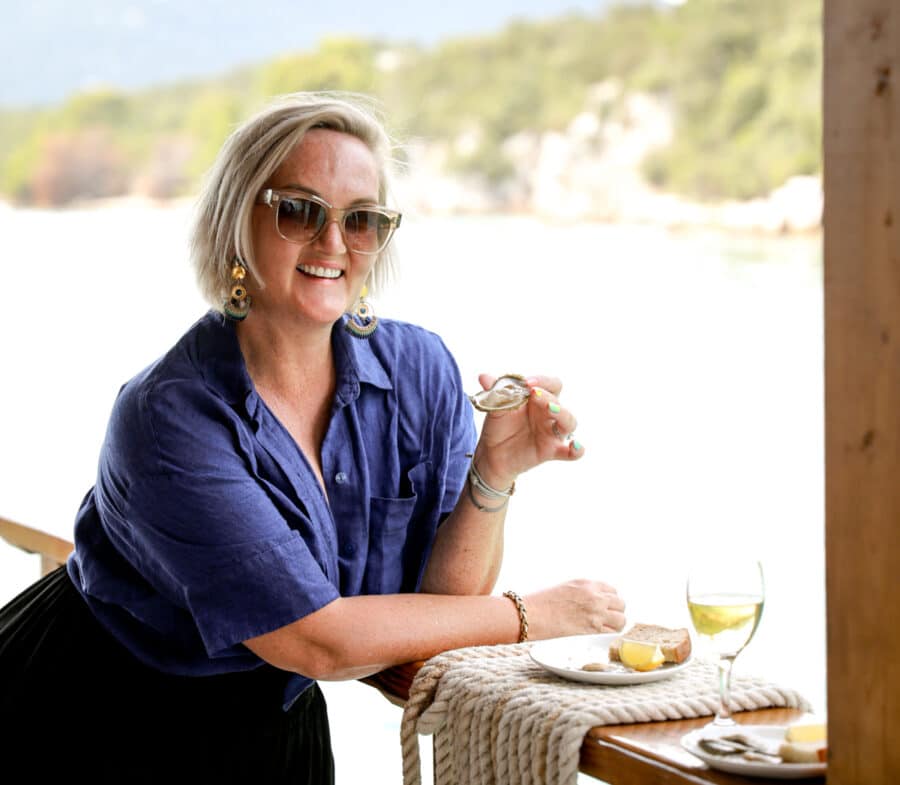 A woman with short blonde hair and sunglasses enjoys oysters and a glass of wine at an outdoor table in Bota Sare, Ston during her picturesque Croatia day trip.