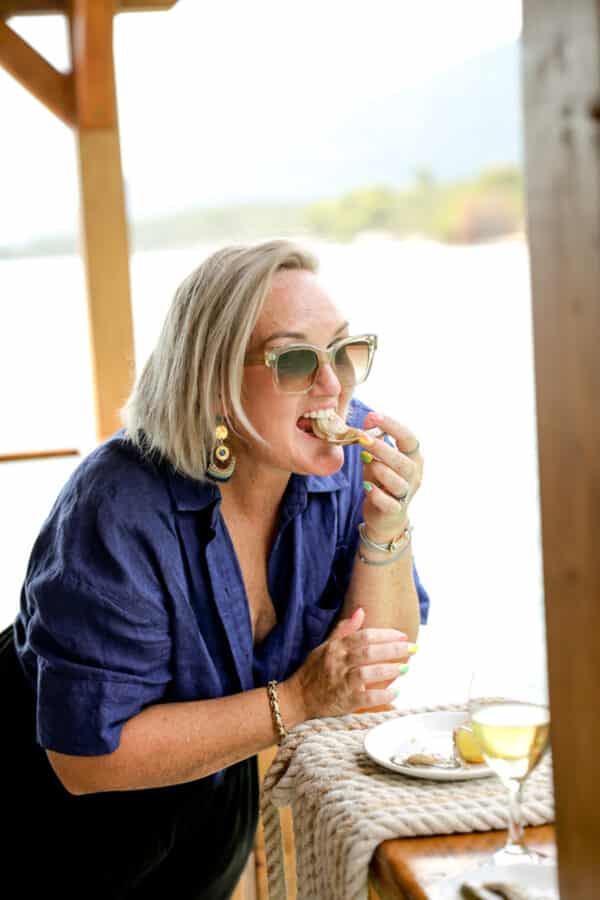 A person with short blonde hair, wearing sunglasses and a blue shirt, enjoys oysters on a veranda near the water. A glass of white wine is on the table. It's the perfect day trip to Bota Sare in Croatia to celebrate Oyster Day.