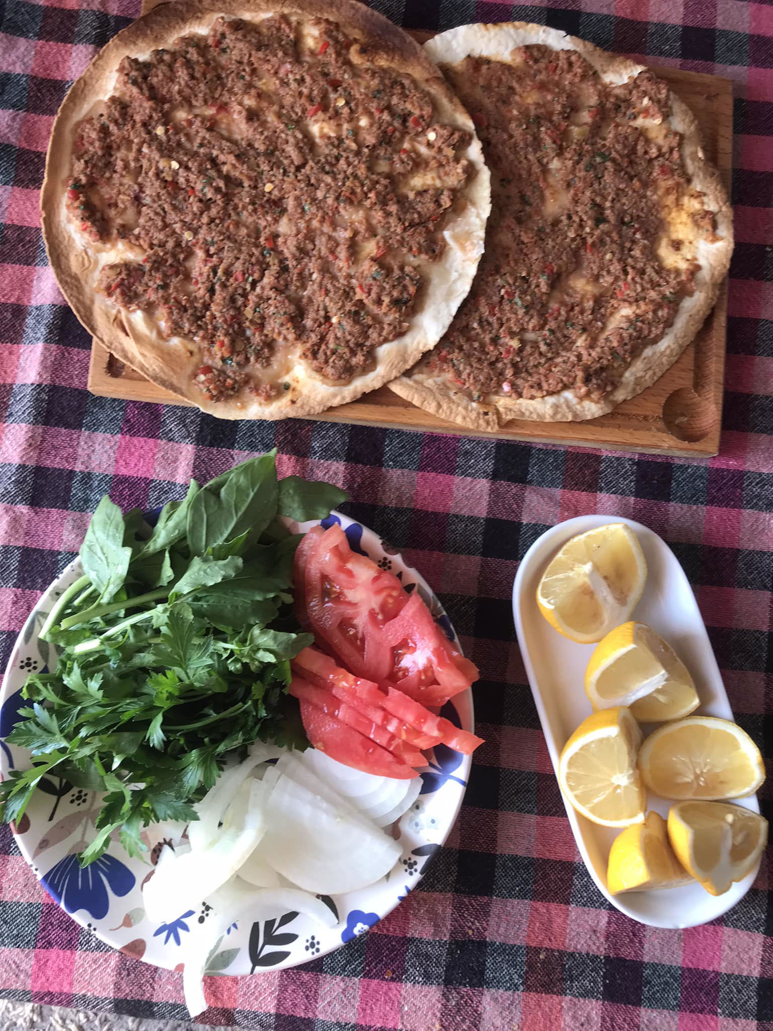 Two Lahmacun with a minced meat topping on a wooden board, alongside a plate of herbs, onions, and tomato slices, and a separate dish with lemon wedges, all set on a checkered tablecloth.