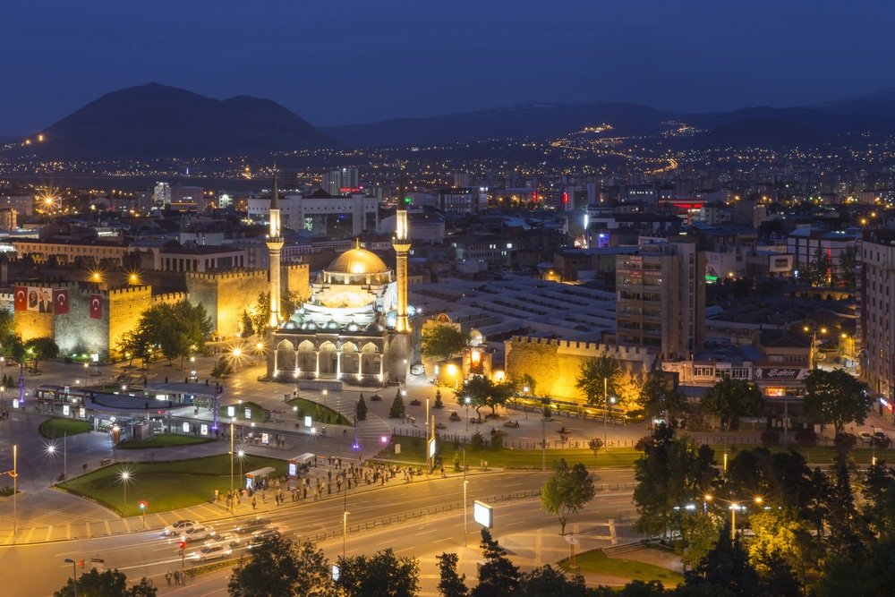 Aerial view of Kayseri city at night featuring a prominently lit mosque with twin minarets, surrounding buildings, and distant mountains in the background, capturing the essence of one of the beautiful cities in Turkey.