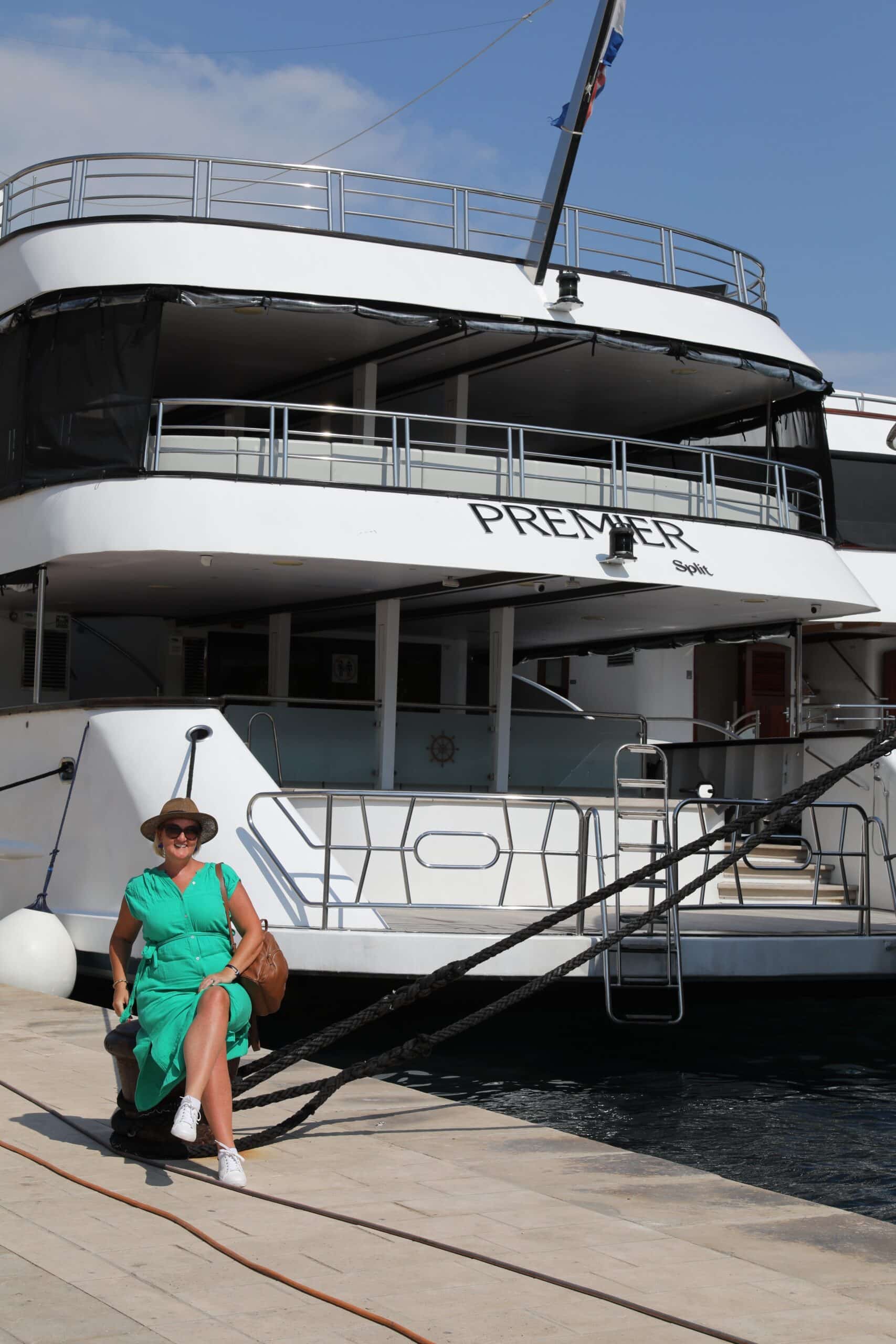 SJ in a green dress sits on a dock beside a large white yacht named 