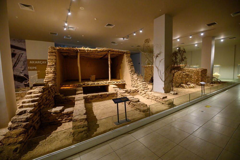 An indoor exhibit at the Sanlıurfa Archaeology Museum showcases a reconstructed ancient dwelling with stone walls and a wooden roof, featuring information displays. This tourist attraction is among the must-see sights for history enthusiasts.