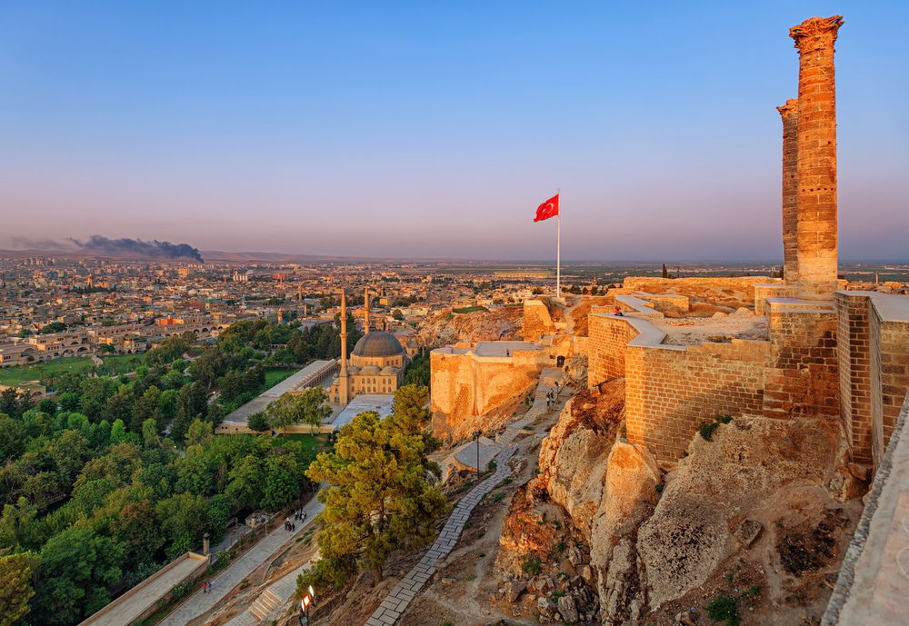 View of the old castle, Urfa, Turkeya with an intact column and Turkish flag overlooking a cityscape at sunset. This popular tourist attraction features green trees and buildings visible below, while smoke rises in the background, adding a touch of mystique to the sights.