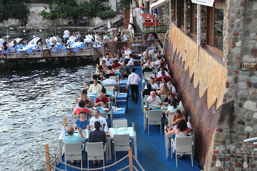 Outdoor waterfront restaurant with diners seated at tables along a dock, under string lights. The restaurant, aptly named Turkish Paradise, is built into the side of a stone building, offering a breathtaking view of the water—a must-visit for travel enthusiasts.