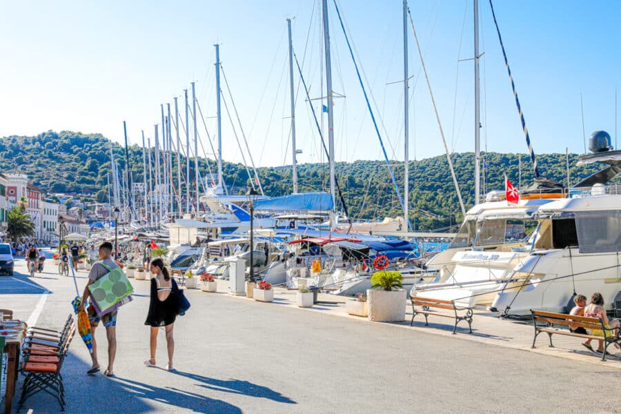 Two women walk along a marina with docked sailboats and yachts in Dubrovnik, carrying beach gear. A sunny, clear day makes the water and sky appear bright blue. Buildings and hills are visible in the background.