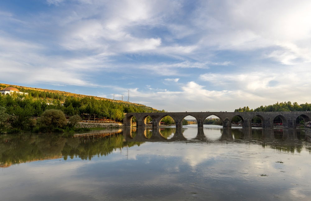 Ten-Eyed Bridge (Ongozlu Bridge) reflected in a calm river, surrounded by lush greenery under a partly cloudy sky, one of the must-see things to do in Diyarbakir.