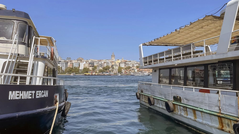 Two boats are docked with a cityscape featuring a prominent tower in the background under a clear blue sky. The boat on the left, named "MEHMET ERCAN," adds to the charm of this picturesque scene showcasing English in Turkey.