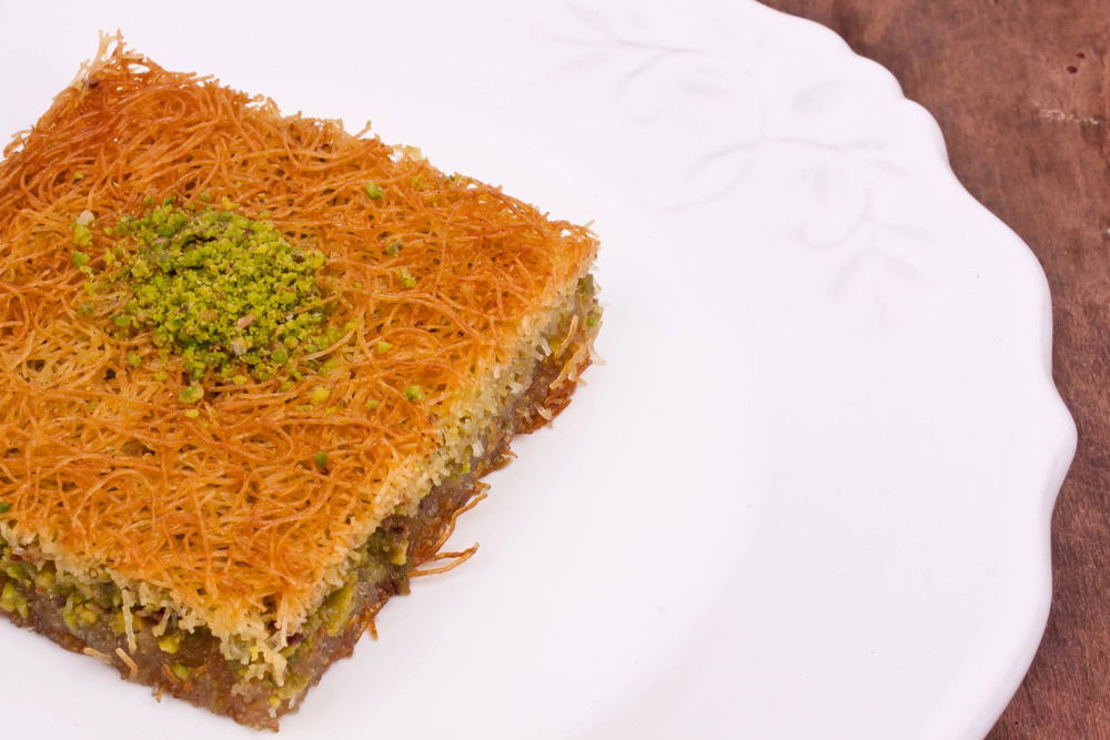 A slice of pistachio Kadayif on a white plate with a detailed edge, showcasing a golden-brown top layer—a must-try delicacy for any Diyarbakir travel guide.