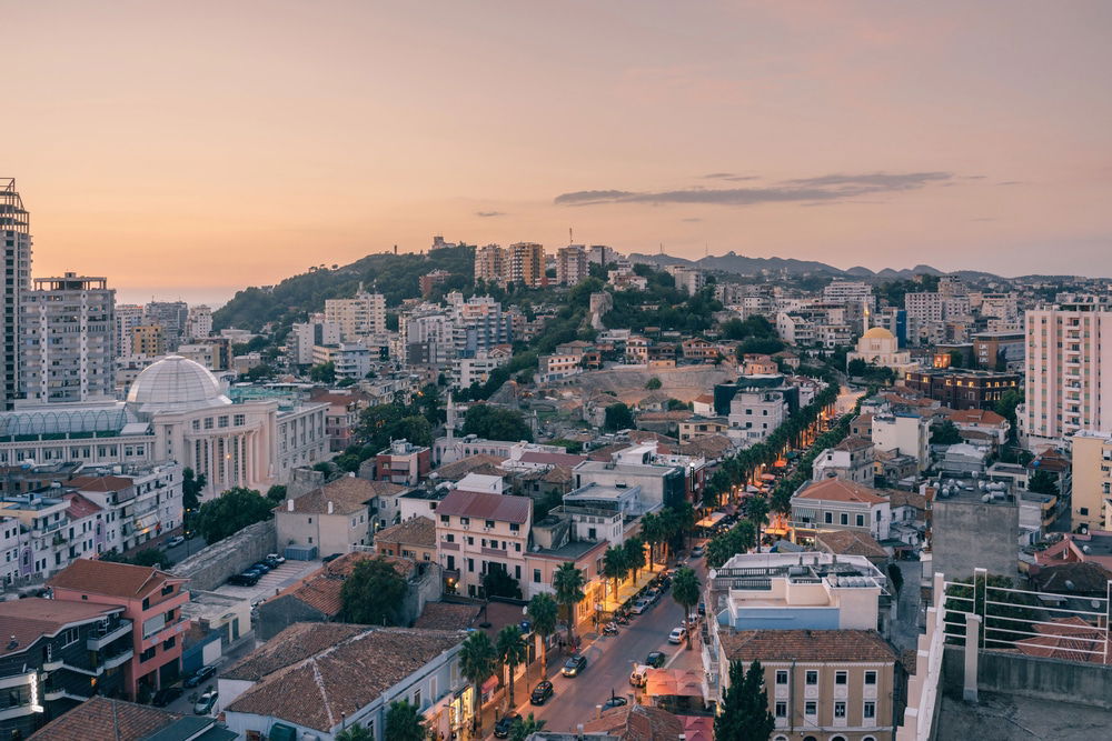 Is Durrës worth visiting? An aerial view of its cityscape at sunset offers a stunning mix of modern and traditional buildings, set against a hillside backdrop, with roads lined in vibrant greenery.