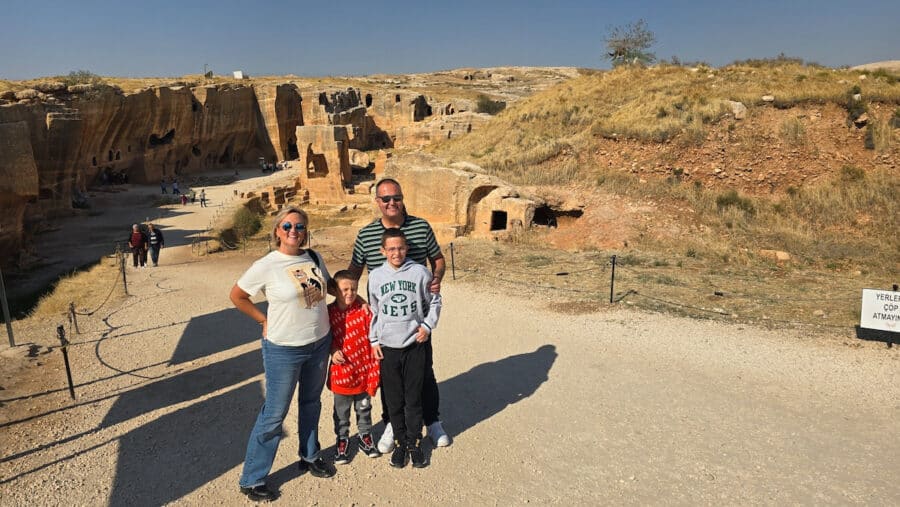 A family of four poses on a path by ancient rock ruins in the Ancient City of Dara, with Mardin, Turkey's Mesopotamian heritage as their backdrop under a clear blue sky.