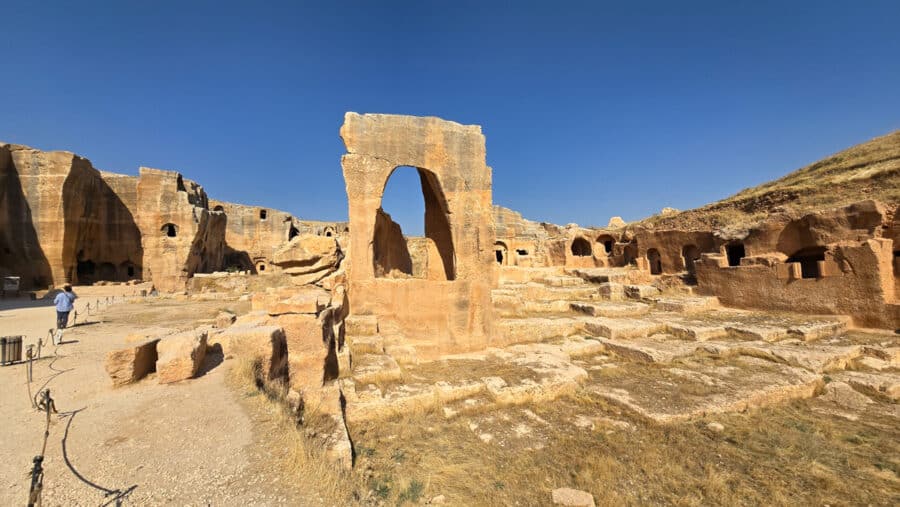 Ancient stone ruins with arches and pathways under a clear blue sky evoke the rich Mesopotamian heritage of Dara. One person walks on a gravel path at the left, exploring the echoes of Mardin's storied past.