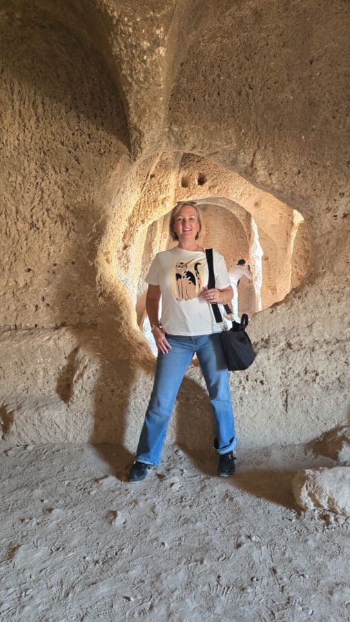 In the Ancient City of Dara, near Mardin, Turkey, a person stands inside a cave-like structure with textured stone walls. They wear a white t-shirt and jeans and carry a black bag as light filters through openings behind them, immersing them in rich Mesopotamian heritage.