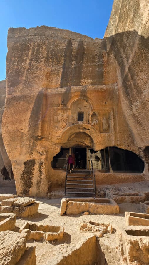 The entrance of an ancient rock-cut structure in the Ancient City of Dara features intricate carvings, with steps leading inside. Sunlit stones surround the doorway, echoing Mesopotamian heritage.