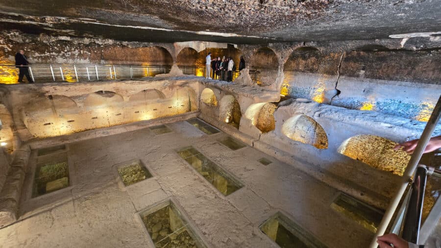 Visitors explore an ancient underground structure in Dara, Mardin, Turkey, with stone arches illuminated by warm lights and glass panels on the floor. This site, rich in Mesopotamian heritage, offers a captivating glimpse into a bygone era.