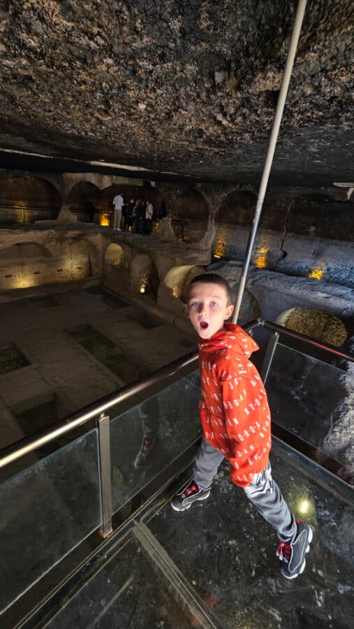 A child in a red jacket stands on a glass walkway inside a dimly lit, ancient stone cave, reminiscent of the majestic Mesopotamian heritage found in the ancient city of Dara near Mardin, Turkey.